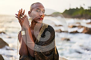 Epatage lgbtq black man poses on scenic ocean beach looks at camera demonstrates jewellery . Androgynous ethnic fashion