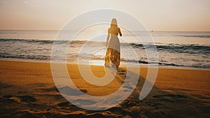 Epic back view shot of young happy woman in summer dress walking to water edge on amazing golden sunset ocean beach.