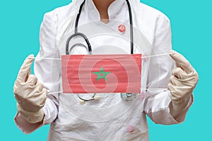 Epidemic in Morocco. Young woman doctor in a medical coat suit and gloves holds a medical mask with the print of the flag of