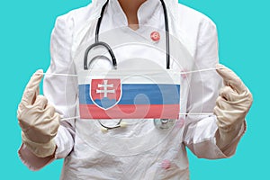 Epidemie na Slovensku. Mladá lékařka v lékařském plášti a rukavicích drží lékařskou masku s potiskem vlajky Slovenska