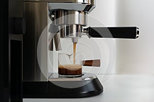 The espresso coffee flowing from coffee machine into a small glass