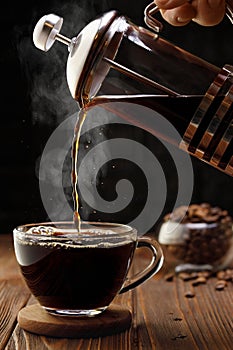 espresso pouring from french press coffee maker into glass cup on wooden table with black background