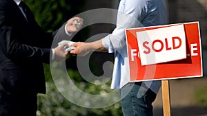 Estate agent and man exchanging money and house keys against sold signboard