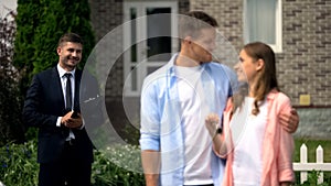 Estate broker watching happy couple enjoying new house showing keys to camera