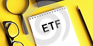 ETF Exchange Traded Funds word written on a notepad next to glasses, a pencil and a magnifying glass on a yellow background