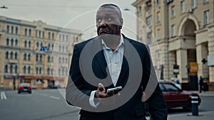 Ethnic adult man African middle-aged businessman entrepreneur with phone in city booking taxi with mobile app waiting