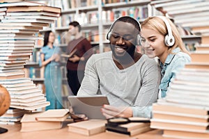 Ethnic african american guy and white girl surrounded by books in library. Students are using tablet.