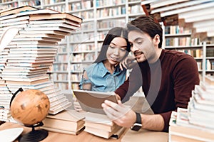 Ethnic asian girl and white guy surrounded by books in library. Students are using tablet.