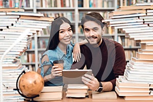 Ethnic asian girl and white guy surrounded by books in library. Students are using tablet.