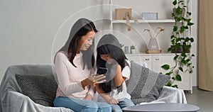 Ethnic family asian mom with daughter watching movie cartoon funny video laughing using mobile phone take selfie on