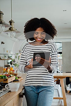 Ethnic female scrolling on digital tablet standing against wooden counter in modern kitchen.