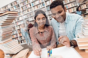 Ethnic indian mixed race girl and guy surrounded by books in library. Students are taking notes.