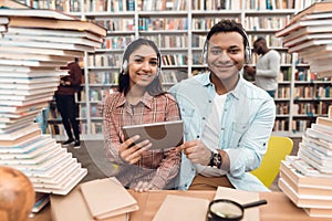Ethnic indian mixed race girl and guy surrounded by books in library. Students are using tablet.