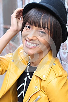 Ethnic woman with toothy smile touching hat and wearing yellow leather jacket