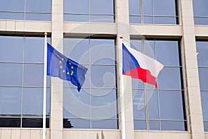 EU flag and Czech Republic flag on flagpole on modern consulate building. Flag of European Union and Czech flag on glass blue