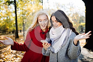 Euphoric friends watching videos on a smartphone and pointing at screen surprised in autumn park