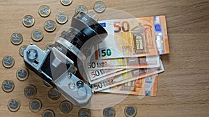 Euro banknotes, coins and a camera on a light wooden background.