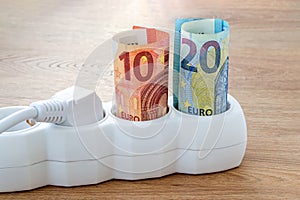 Euro banknotes and white power plug inserted into an extension socket on a wooden surface. Expensive energy and cost of