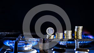 A euro coin along with a row of yellow coins on a gas stove against a blue flame of natural gas on a dark background