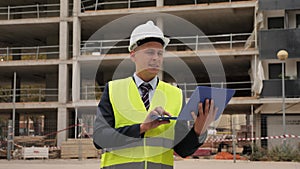 European architect in workwear and white hardhat with laptop in hands standing on building site.