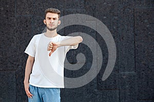 European bearded scowling man in white T-shirt and jeans showing thumbs down dislike with his left hand outside