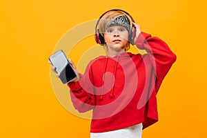 European blond boy in a red hoodie listens to music in red headphones and holds smartphone with mockup on a orange