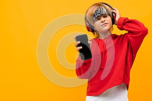 European blond boy in a red hoodie listens to music in red headphones and holds smartphone on a yellow background