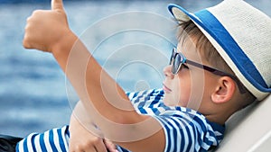 European boy in sunglasses and hat lying on deck chair relaxing with crossed arms showing thumb up