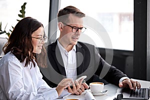 European Business Lady And Manager Discussing Projects And Ideas, Using Smartphone And Laptop, Over Cup Of Coffee