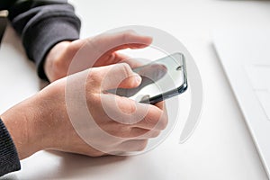 European child hands holding black smartphone with fingers and black screen on white desk infront of white notebook at school