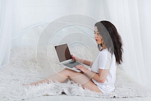 European girl with dark in white t-shirt is using a laptop sitting on the bed.