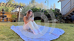 European Girl is sitting in a clearing on a white blanket in white clothes in her hands with a phone. She is looking at