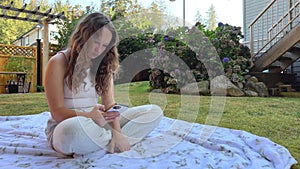 European Girl is sitting in a clearing on a white blanket in white clothes in her hands with a phone. She is looking at