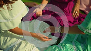 European girls friends in dresses sitting in a circle holding each other`s hands in nature with green grass on the background