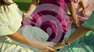 European girls friends in dresses sitting in a circle holding each other`s hands in nature with green grass on the background