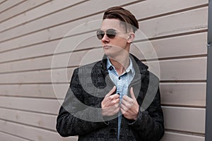 European hipster young man in a trendy plaid jacket in a vintage shirt in fashionable sunglasses with a stylish hairstyle poses