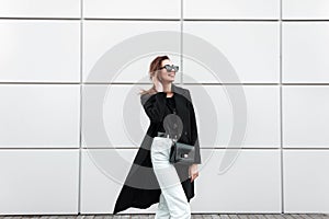 European joyful young hipster woman in a vintage black long coat in white jeans with a stylish leather handbag in dark sunglasses