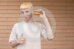 A European man dressed in a beige T-shirt holding a burger in hand near the face, showing thumbs up