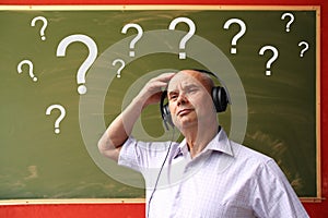 European man in headphones clings to his head amid a lot of white question marks on a chalk board for chalk, text, concept close-