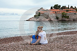 European mom and daughter sitting together on the luxury beach against Sveti Stefan hotel and resort. Adriatic sea, Montenegro