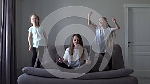 European mother meditating sitting on sofa while active energetic two kids daughters jumping playing in modern home