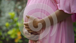 European pregnant girl in a pink dress gently touches her big stomach with her hands, stands on the back green garden