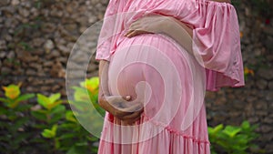 European pregnant girl in a pink dress gently touches her big stomach with her hands, stands on the back green garden