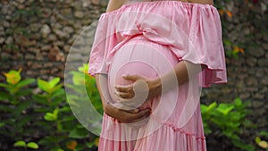 European pregnant girl in a pink dress gently touches her big stomach with her hands, stands on the back green garden