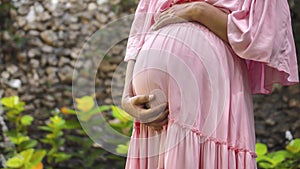 European pregnant girl in a pink dress gently touches her big stomach with her hands, stands on the back green garden