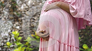 European pregnant girl in a pink dress gently touches her big stomach with her hands, stands on the back green garden