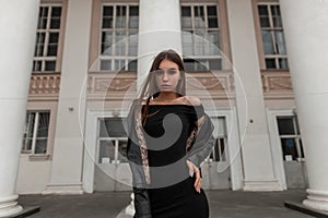 European sexy young woman in an elegant black dress in a fashionable leather jacket with brown hair posing in the city near a