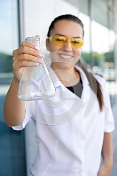 European smiling female scientist in protective coat and yellow glasses shows empty Erlenmeyer flask