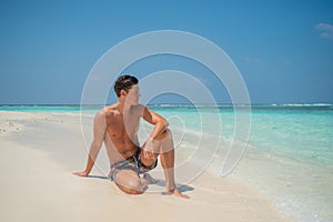 European tanned man wearing swimming shorts sitting at tropical sandy beach at island luxury resort
