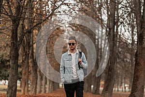 European trendy handsome young man in stylish jeans clothes in vintage sunglasses with a fashionable hairstyle with backpack walks